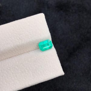 Puede incluir: Una vibrante esmeralda de corte rectangular. La gema es de un verde brillante y transl&uacute;cido, y descansa sobre una superficie blanca y texturizada. El fondo es de un color oscuro y s&oacute;lido, que contrasta con la gema.