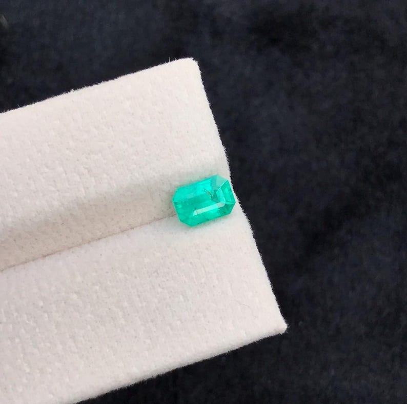 Puede incluir: Una vibrante esmeralda de corte rectangular. La gema es de un verde brillante y transl&uacute;cido, y descansa sobre una superficie blanca y texturizada. El fondo es de un color oscuro y s&oacute;lido, que contrasta con la gema.