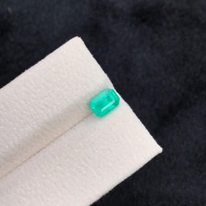 Puede incluir: Una esmeralda verde vibrante, tallada en forma rectangular. La gema se muestra sobre una tela blanca doblada, contra un fondo oscuro. Las facetas de la esmeralda reflejan la luz, mostrando su claridad y color.