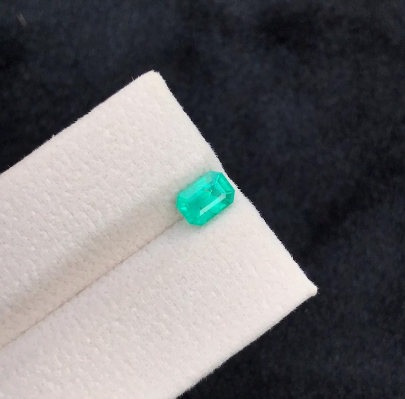 Puede incluir: Una esmeralda verde vibrante, tallada en forma rectangular. La gema se muestra sobre una tela blanca doblada, contra un fondo oscuro. Las facetas de la esmeralda reflejan la luz, mostrando su claridad y color.