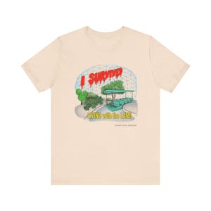 Op de afbeelding: Crèmekleurig T-shirt met een afbeelding van een kas met een boottocht en de tekst "I Survived Living with the Land". De tekst is rood en groen, met de merknaam onderaan.