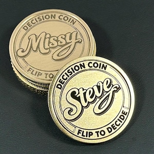 May include: Gold-colored decision coins with the names "Missy" and "Steve" engraved on them. Each coin has the words "Decision Coin" and "Flip to Decide" around the edges. The coins are stacked on a dark surface.