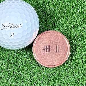 May include: A Titleist golf ball with the number 2 next to a copper-colored golf ball marker with the number 11 in tally marks. The items are on a green artificial turf surface.