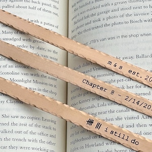 Può includere: Tre segnalibri in legno con bordi strutturati sono appoggiati su un libro. Ogni segnalibro ha il testo stampato: "m&s est. 2018", "Chapter 8 - 2/14/2018" e "i still do". I segnalibri sono di colore marrone chiaro.