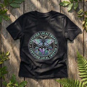 Puede incluir: Camiseta negra con un diseño gráfico circular que presenta una polilla, setas y cristales. El texto "CERTIFIED LOCAL CRYPTID" rodea el diseño en color turquesa. La polilla tiene alas azules, verdes y moradas.