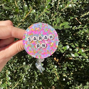 Peut inclure: Un porte-badge rond et scintillant avec le nom "SARAH RN" épelé avec des perles de lettres blanches. Le porte-badge contient un mélange de paillettes roses, bleues et jaunes. L'arrière-plan est un buisson vert.