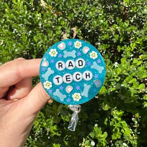 May include: A round, teal badge reel with the words "RAD TECH" spelled out in white letter beads. The badge features bone, flower, and rainbow designs. The reel is held by a hand against a background of green foliage.