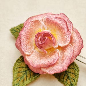 May include: A detailed embroidered pink rose with a yellow centre and green leaves. The flower is crafted with fine stitching, creating a realistic design. Likely a brooch or hair accessory.