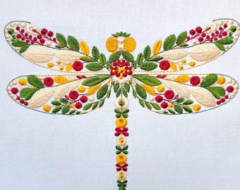 May include: An embroidery design of a dragonfly on a white fabric background. The dragonfly features wings filled with green leaves, red berries, yellow petals, and cream-colored leaf shapes. The body is composed of stacked yellow and red floral motifs.