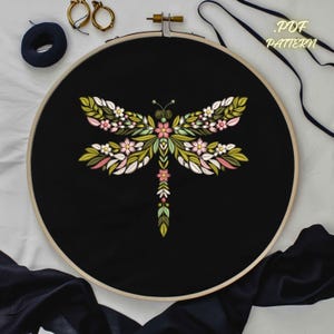 May include: A black embroidery hoop featuring a floral dragonfly design. The dragonfly is embroidered with pink, white, and green thread. The text "PDF PATTERN" is visible on the upper right. Additional embroidery supplies are visible.