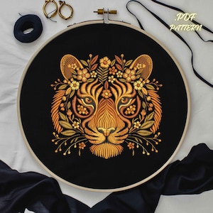 May include: Embroidered tiger face on black fabric, framed in a wooden hoop. The tiger design features intricate details in shades of gold and orange, with floral accents. The text "PDF PATTERN" is visible.