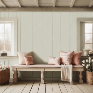 May include: A white bench with a light pink cushion and pink throw pillows sits in front of a light green wall. The bench has four legs and a wooden frame. The wall is made of wood planks. There are two windows on either side of the bench.