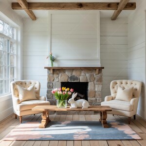 May include: A living room with a stone fireplace, two white armchairs, a wooden coffee table, a white rabbit figurine, and a bouquet of pink and yellow flowers in a glass vase. The room has white walls and a wooden beam ceiling.
