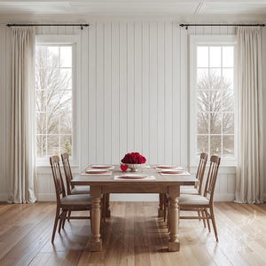 May include: A dining room with a wooden table set for a meal. The table is surrounded by wooden chairs and set with plates and placemats. A centerpiece of red roses sits in a silver bowl. Two windows with sheer curtains flank the table.