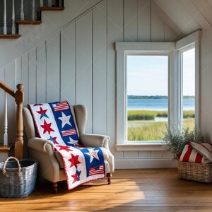Puede incluir: Un interior acogedor con un sillón color crema adornado con un edredón patriótico. El edredón presenta estrellas rojas, estrellas blancas y rayas sobre un fondo azul y blanco. Una cesta de mimbre y una ventana con vistas al agua completan la escena.