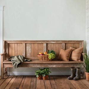 May include: A rustic wooden bench with a woven basket filled with fruit and greenery. Two brown throw pillows are on the bench, and a green jacket is draped over the seat. Two brown boots are on the floor next to the bench, and two potted plants are in front of the bench.