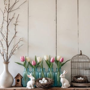 May include: A white wooden background with a row of blue glass bottles filled with pink and white tulips.  Two white ceramic bunnies are on either side of the bottles.  A birdcage with a nest of white eggs is on the right side of the image.