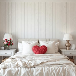 May include: A bedroom scene with a bed featuring white pillows and a red heart-shaped cushion. Two bedside tables hold lamps, flowers, and books. The wall is panelled in white wood.