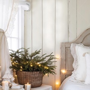 May include: A wicker basket holding evergreen branches and fairy lights. The basket is on a white table beside a bed with a distressed headboard and white bedding. A window with lace curtains is in view.