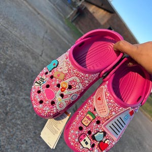 Pode incluir: Crocs cor-de-rosa decoradas com pingentes temáticos médicos, incluindo um estetoscopio, bandagens, pílulas e uma máscara facial. Os pingentes são cercados por strass rosa e branco.