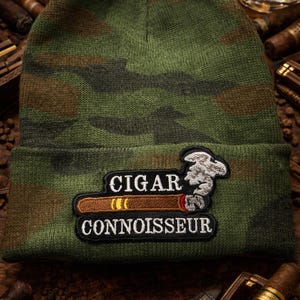 Op de afbeelding: Groene camouflage beanie met een patch met de tekst "CIGAR CONNOISSEUR" boven een afbeelding van een brandende sigaar. De beanie is omgeven door sigaren en een glas amberkleurige vloeistof.