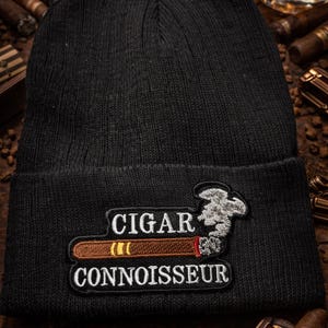 Op de afbeelding: Zwarte gebreide muts met een patch met de tekst "CIGAR CONNOISSEUR". De patch toont een bruine sigaar met een rode punt en een rookwolk. De muts is donker van kleur en lijkt van zacht materiaal te zijn gemaakt.