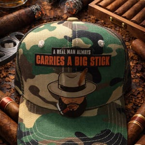 May include: A camouflage baseball cap featuring a rectangular patch with the text A REAL MAN ALWAYS CARRIES A BIG STICK and an illustration of a person wearing a fedora with a feather and smoking a cigar, with white stitching on the brim.