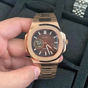 May include: A rose gold Seiko automatic watch with a brown dial and a visible escapement. The watch features a square case with a rounded bezel and a matching rose gold bracelet. The watch face has white hour markers and hands.