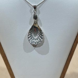 Op de afbeelding: Een zilveren ketting met een schelp hanger op een houten standaard.