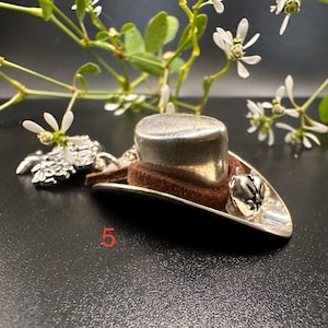 Peut inclure: Un petit charme en forme de chapeau haut-de-forme argenté avec une bande marron, fixé à une base argentée. Une petite cloche argentée et le chiffre "5" en rouge sont également visibles. Le charme est posé sur un fond sombre avec des fleurs blanches.