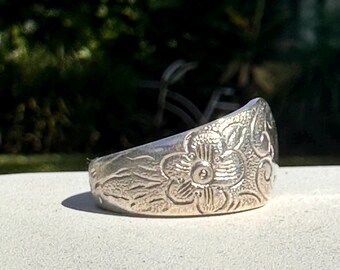 May include: A silver ring with a floral design. The ring is wide and has a curved shape. The flower design is intricate and detailed.