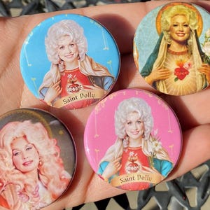 May include: Four round buttons with images of a woman with blonde hair wearing a blue and white dress and holding a heart. The buttons have a pink, blue, and white background with the text "Saint Dolly" on each button.