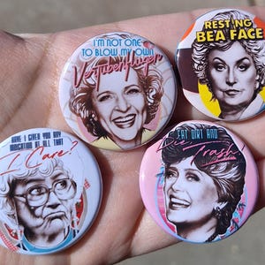 Può includere: Quattro spille rotonde con ritratti di donne e frasi umoristiche. Le spille hanno sfondi colorati e frasi come "I'm not one to blow my own Verbenfluffer" e "Resting Bea Face".