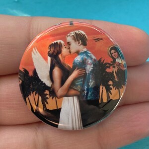 May include: A round button with a colorful image of a couple kissing in front of a sunset. The couple is surrounded by palm trees and the woman has angel wings. There is a small image of a woman in the upper right corner.