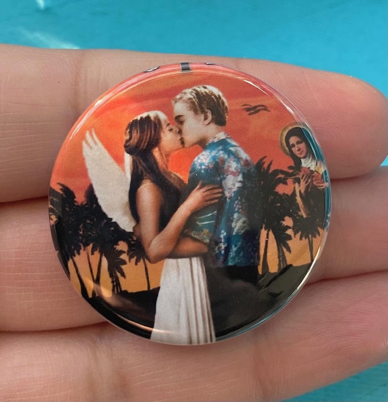 May include: A round button with a colorful image of a couple kissing in front of a sunset. The couple is surrounded by palm trees and the woman has angel wings. There is a small image of a woman in the upper right corner.
