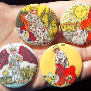 Puede incluir: Cuatro botones redondos de cartas del tarot. Cada botón presenta una ilustración diferente con colores vibrantes. Los botones representan figuras, símbolos y escenas asociadas con las imágenes de las cartas del tarot, incluyendo una mujer, un sol, un ángel y una mujer con un león.