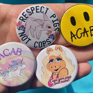 May include: Four round button pins with different designs. The first pin is pink and white with a pig and the text "Respect Not Cops Pigs". The second pin is yellow with a smiley face and the text "ACAB". The third pin is pink and white with a unicorn and the text "ACAB". The fourth pin is pink and white with a pig and the text "Cops give pigs a bad name!"