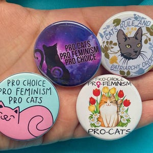 Puede incluir: Cuatro pines redondos con lemas a favor de los gatos y el feminismo. Los pines presentan un fondo de galaxia, un gato gris, un gato rosa y un gato naranja de dibujos animados con flores.
