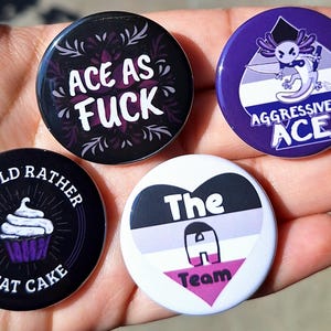 May include: Four circular badges with different designs. One says "ACE AS FUCK" in white on black. Another features an axolotl and the words "AGGRESSIVELY ACE". The others say "I WOULD RATHER EAT CAKE" and "The A Team".
