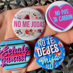 May include: Four round button pins with colorful designs and Spanish phrases. The pins feature a floral design with the phrase "No me jodas", a blue and white striped design with the phrase "Échale ganas", a blue design with cherries and the phrase "No te dejes mija", and a pink heart with the phrase "No pos ta cabron".