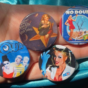 May include: Four round blue and white buttons with band names and album art. The buttons feature the bands "Aqua", "No Doubt", "Mellon Collie and the Infinite Sadness", and a band with a woman in a white dress.