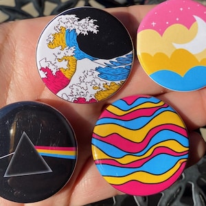 Peut inclure: Quatre badges ronds avec des motifs différents. Le premier badge représente une vague bleue et jaune avec une montagne rose en arrière-plan. Le deuxième badge représente un ciel rose avec des nuages jaunes et une lune croissante blanche. Le troisième badge représente un triangle noir avec une bande arc-en-ciel. Le quatrième badge représente un motif ondulé bleu, jaune et rose.