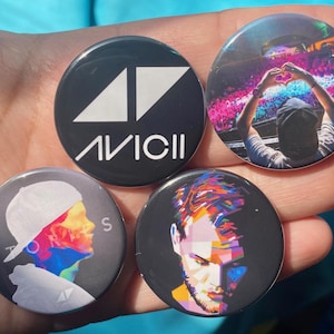 Puede incluir: Cuatro botones redondos con diferentes diseños. El primer botón es negro con un logotipo triangular blanco y el texto "AVICII". El segundo botón es negro con un retrato colorido de un hombre. El tercer botón es negro con un retrato colorido de un hombre con un sombrero blanco. El cuarto botón es negro con una imagen colorida de una persona con los brazos en alto.