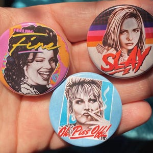 May include: Three round button badges with colourful designs and images of women. The first badge features a woman with long curly hair and the text "Feeling Fine". The second badge features a woman with long straight hair and the text "SLAY". The third badge features a woman with short blonde hair and the text "Oh Piss Off!"