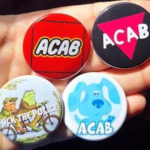 May include: Four circular badges with different designs. One has "ACAB" in white on a red background, another has "ACAB" in white on a black background with a pink triangle. The other two badges feature cartoon characters and text.