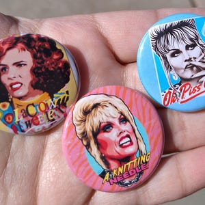 May include: Three round pinback buttons featuring portraits of women. One button has the text "La Croix Sweets", another says "Oh Piss Off!", and the third reads "A Knitting Needle!"