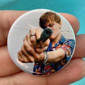 May include: A close-up of a man holding a gun, aiming it directly at the camera. He is wearing a colorful, patterned shirt and has a serious expression on his face.