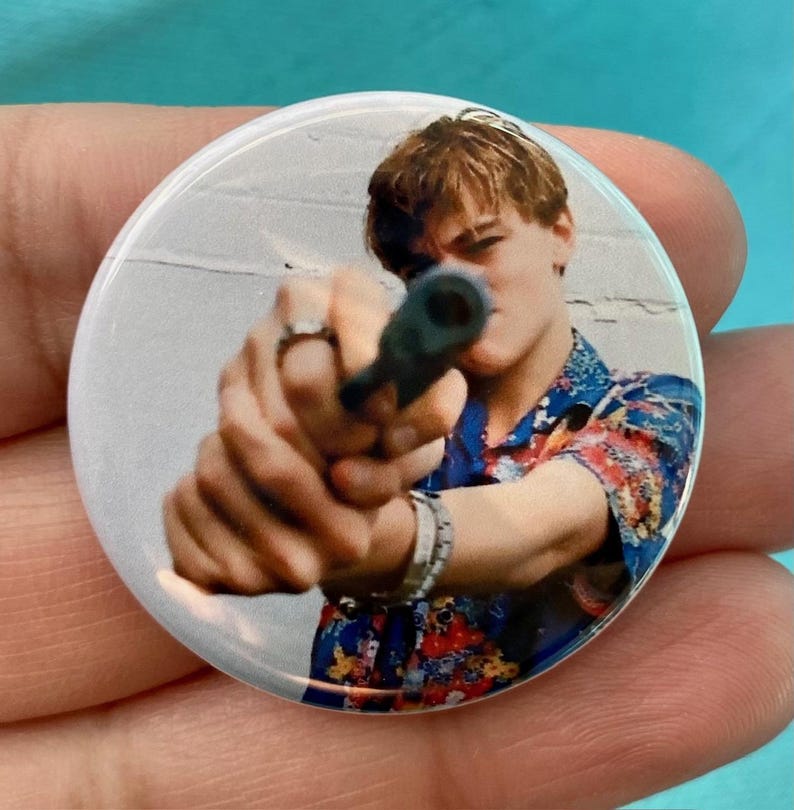 May include: A close-up of a man holding a gun, aiming it directly at the camera. He is wearing a colorful, patterned shirt and has a serious expression on his face.