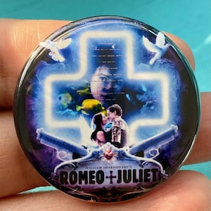 May include: A round button with a blue and purple background featuring a white cross with a couple in the center. The couple is standing in front of a blue and purple background with fish and doves. The text "ROMEO + JULIET" is at the bottom of the button.