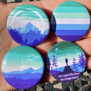 May include: Four round pins with a blue, green and white colour scheme. The pins feature a mountain range, a sky with birds, a forest scene with a person standing on a rock, and a flag with five horizontal stripes. The text on the forest scene pin reads "love opens the doors we've kept closed."